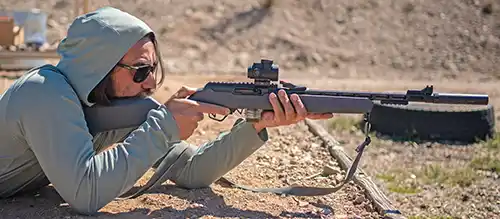 Shooter aiming a Stealth Grey TandemKross TKX22 Light Rifle with a Trijicon red dot from prone at an outdoor range.